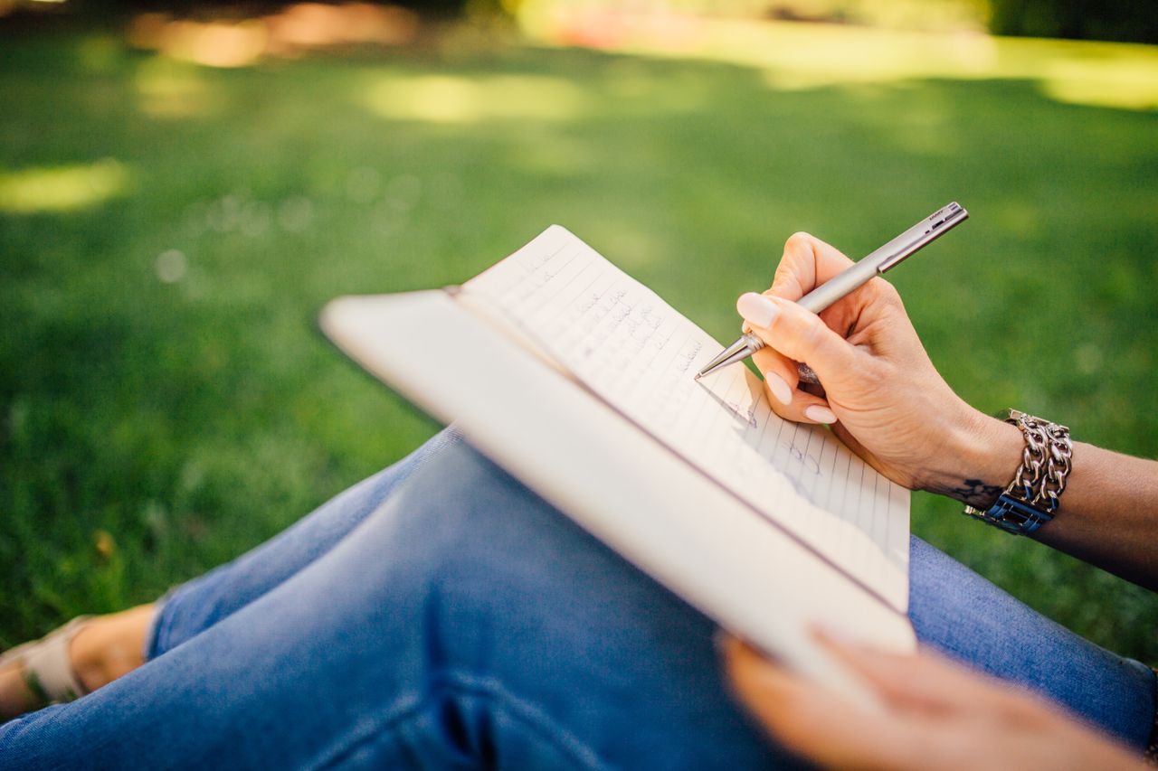 About Person writes in a notebook on a sunny day. Focus on hands and pen outdoors.